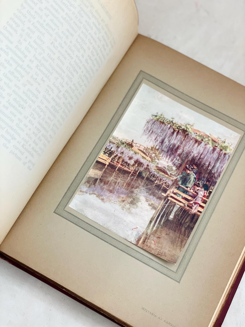 May include: An open book with a framed watercolor print of a Japanese scene. The print depicts a wooden bridge over water, with people and wisteria flowers. The book's pages are aged, and the print is titled "Wistaria at Kameda".