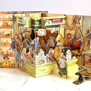 May include: A multi-layered pop-up book featuring a detailed toy shop scene. The illustration includes various figures such as a jester, a person sitting on a counter, an elephant, a monkey, and a white dog, all rendered in a colorful, artistic style.