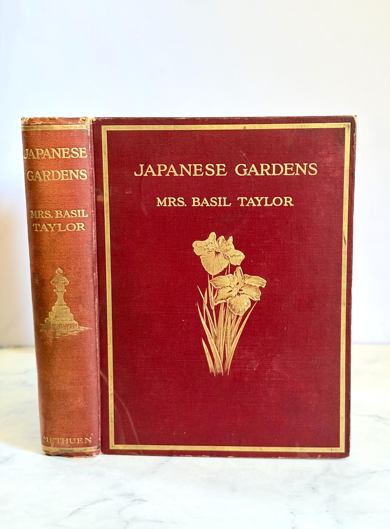 May include: Two antique books titled "Japanese Gardens" by Mrs. Basil Taylor. The books have a red cover with gold lettering and a gold floral design. The spine of the book on the left is also red with gold lettering.