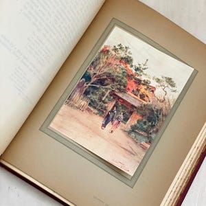 May include: An open book displays a watercolor print of a Japanese garden scene. The print features two figures walking towards a gate, with trees and foliage in the background. The book's pages are aged, and the print is framed.