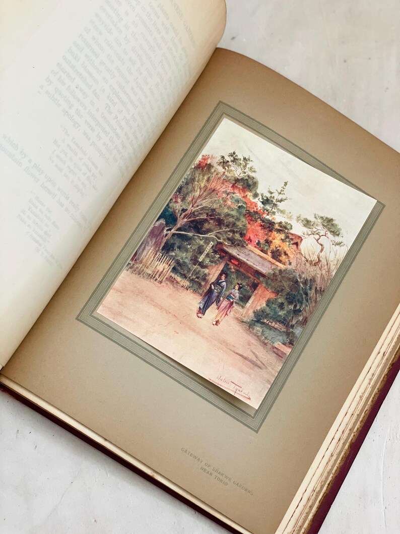 May include: An open book displays a watercolor print of a Japanese garden scene. The print features two figures walking towards a gate, with trees and foliage in the background. The book's pages are aged, and the print is framed.