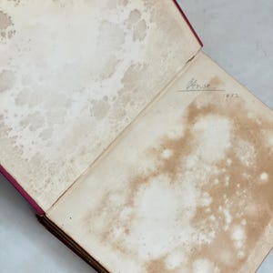May include: An open antique book with aged, stained pages. The pages show water damage and discoloration, with a handwritten inscription and the year 1912 visible. The book's cover is a deep red.