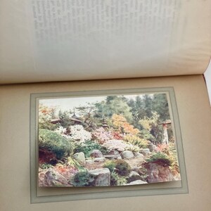 May include: A photograph of a Japanese rock garden at Nikko, featuring a variety of colorful plants and flowers. The garden includes large rocks, a pathway, and a traditional Japanese structure in the background. The image is framed within a vintage photo album.