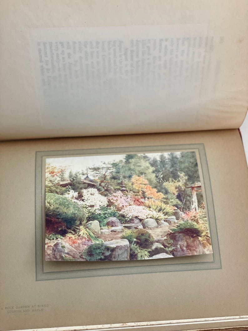 May include: A photograph of a Japanese rock garden at Nikko, featuring a variety of colorful plants and flowers. The garden includes large rocks, a pathway, and a traditional Japanese structure in the background. The image is framed within a vintage photo album.