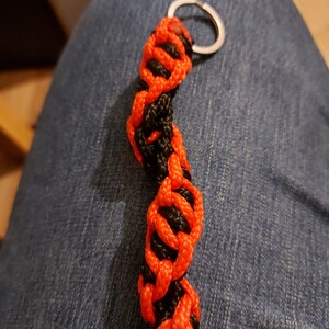 May include: A handmade keychain featuring a braided design in black and bright orange. The keychain is made of woven cord and has a silver-colored metal ring for attaching keys. The design is a repeating pattern of interwoven strands.