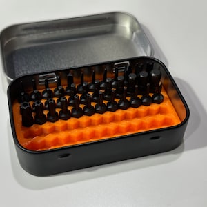 Puede incluir: Una caja de metal negro con un inserto de espuma naranja contiene una colección de pequeños bits de destornillador negros.