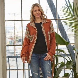 May include: A rust-colored, long-sleeved shirt with a leopard print design. The shirt has a button-up front and a relaxed fit. The model is wearing the shirt with blue jeans and black ankle boots.