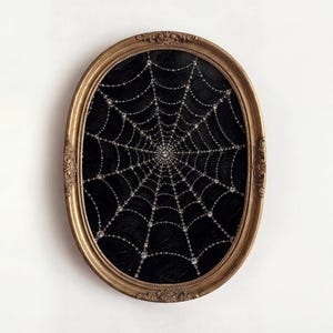 Op de afbeelding: Een ovaalvormig wandkunstwerk met een zwarte achtergrond en een gedetailleerd spinnenwebontwerp bestaande uit kleine, reflecterende kralen of kristallen. Het kunstwerk is gevat in een sierlijke, antieke gouden lijst met decoratief houtsnijwerk.