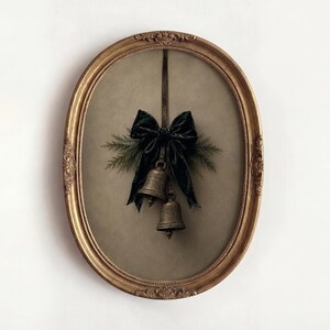 Può includere: Una cornice ovale dorata e decorata contenente una stampa di due campane metalliche appese a un nastro scuro. Un fiocco in velluto verde scuro è legato sopra le campane, accentuato da piccoli rametti di pino verde su uno sfondo neutro.
