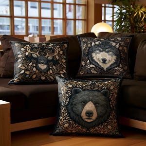 Könnte beinhalten: Drei dekorative Kissen mit Tierporträts auf einem braunen Sofa. Die Kissen zeigen einen Hirsch, einen Wolf und einen Bären, jeweils umgeben von einem floralen Design. Die Kissen sind schwarz mit goldenen und cremefarbenen Akzenten und verleihen dem Raum Eleganz.