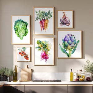Könnte beinhalten: Eine Sammlung gerahmter Aquarelldrucke mit verschiedenen Gemüsesorten. Die Drucke zeigen einen Kopf Salat, Karotten, Knoblauch, Tomaten, Radieschen und eine Artischocke. Die Kunstwerke werden in einer Küche mit neutraler Farbpalette ausgestellt.