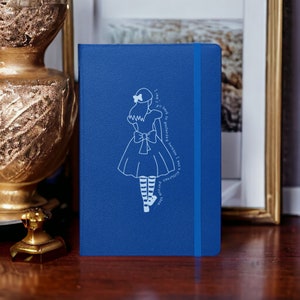 Op de afbeelding: Een blauw notitieboek met een witte illustratie van een meisje in een jurk en de tekst "Ik kan niet terug naar gisteren, omdat ik toen een ander persoon was..."