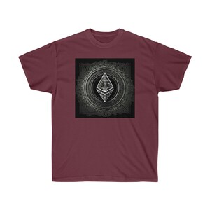 Puede incluir: Una camiseta color granate con un gráfico cuadrado negro que presenta un diseño geométrico blanco que se asemeja al logotipo de la criptomoneda Ethereum. El diseño está rodeado por un patrón circular de líneas y puntos.