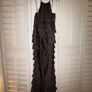May include: A dark brown, floor-length formal dress with a halter neckline and tiered skirt. The dress features a decorative belt with gold-colored embellishments. The dress is hanging on a black velvet hanger against a white shuttered background.
