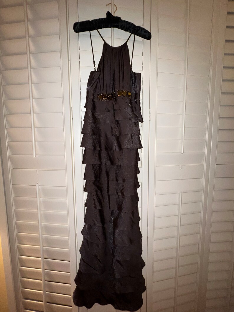 May include: A dark brown, floor-length formal dress with a halter neckline and tiered skirt. The dress features a decorative belt with gold-colored embellishments. The dress is hanging on a black velvet hanger against a white shuttered background.