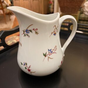 May include: White ceramic pitcher with a curved spout and handle. The pitcher is decorated with small floral sprigs in shades of blue, red, and yellow. The pitcher is sitting on a black surface.
