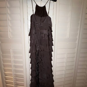 May include: A dark brown, tiered formal dress with spaghetti straps hangs on a black velvet hanger. The dress features a halter neckline and a series of horizontal ruffles cascading down the length of the garment. The dress is made of a shiny fabric.