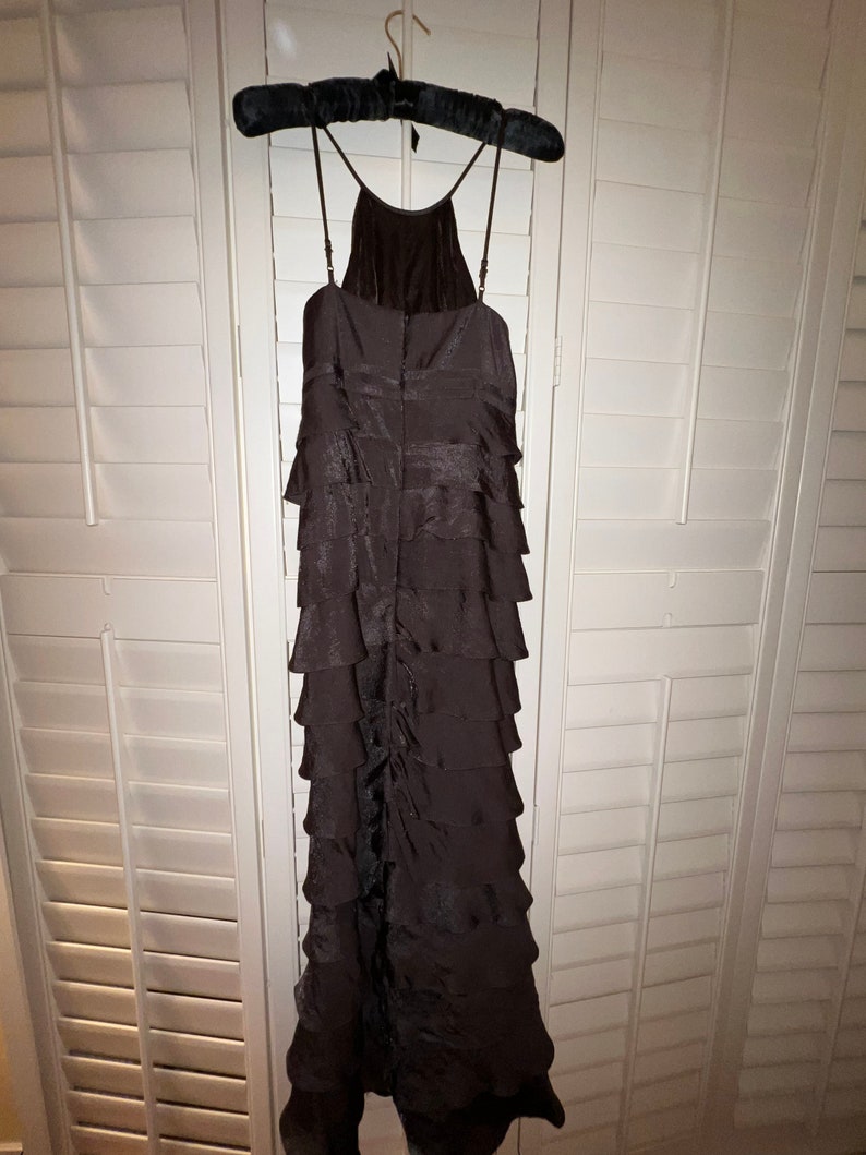 May include: A dark brown, tiered formal dress with spaghetti straps hangs on a black velvet hanger. The dress features a halter neckline and a series of horizontal ruffles cascading down the length of the garment. The dress is made of a shiny fabric.