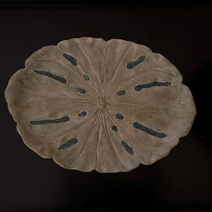 May include: A ceramic platter shaped like a large, flat lotus leaf with a muted sage green finish. The surface features a radial vein pattern and several elongated, textured cutouts in a dark, speckled blue-gray tone radiating from the center.