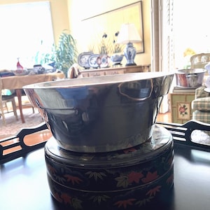 May include: A large, dark blue bowl sits atop a black, round container with a decorative floral pattern in red and green. The bowl has a shiny, reflective surface. The items are on a black tray.