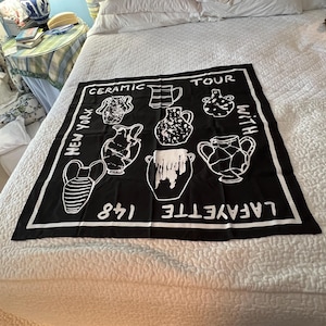 May include: Black square scarf with white illustrations of ceramic vessels. The scarf has the words "Ceramic Tour With New York Lafayette 148" printed in white. The scarf is displayed on a white bedspread.