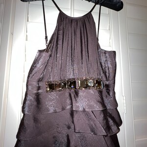 May include: A dark brown, tiered dress with a halter neckline, hanging on a black padded hanger. The dress features a decorative belt with square embellishments. The fabric has a subtle sheen, and the dress appears to be made of a flowing material.