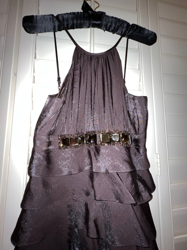 May include: A dark brown, tiered dress with a halter neckline, hanging on a black padded hanger. The dress features a decorative belt with square embellishments. The fabric has a subtle sheen, and the dress appears to be made of a flowing material.