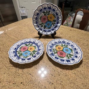 Op de afbeelding: Drie witte keramische borden met geschulpte randen, voorzien van een handgeschilderd bloemmotief in het midden met roze, gele en blauwe bloemen omringd door groene bladeren, en een blauw decoratief randpatroon rondom.
