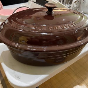 May include: A dark brown Le Creuset Dutch oven with a matching lid. The oval-shaped pot has a rounded base and side handles. The lid features the brand name and a small, round handle. The pot is sitting on a white surface.