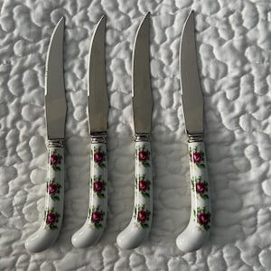 May include: Four steak knives with serrated blades and white handles decorated with red roses and green leaves. The knives have a silver blade and a curved handle design.