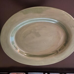 May include: An oval-shaped ceramic serving platter with a smooth, glossy finish in a muted sage green colour. The dish features a slightly raised rim and a shallow centre, showcasing subtle variations in the glaze across its surface.