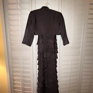 May include: A dark brown formal dress with a matching bolero jacket. The dress features a tiered, ruffled skirt and is hanging on a hanger. The bolero jacket has long sleeves and a high neckline. The outfit is displayed against a white shuttered background.