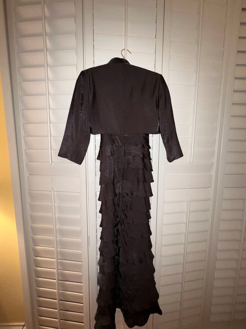 May include: A dark brown formal dress with a matching bolero jacket. The dress features a tiered, ruffled skirt and is hanging on a hanger. The bolero jacket has long sleeves and a high neckline. The outfit is displayed against a white shuttered background.