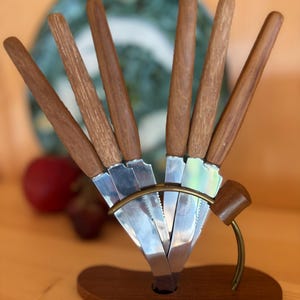 Könnte beinhalten: Ein Set aus sechs Steakmessern mit Holzgriffen und Edelstahlklingen, präsentiert in einem Holzhalter. Die Messer sind fächerförmig angeordnet und werden von einem messingfarbenen Metallring zusammengehalten. Der Halter steht auf einer Holzoberfläche.
