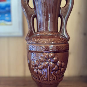 Könnte beinhalten: Eine glänzend braune Keramikvase mit zwei seitlichen Griffen, einem hohen Hals und einem bauchigen Körper. Die Außenseite ist mit einem erhabenen Reliefmuster aus stilisierten Blumen und geometrischen Bändern verziert.