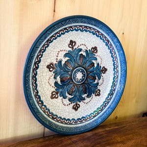 Könnte beinhalten: Ein runder Keramikteller mit weißer Craquelé-Glasur. In der Mitte befindet sich ein blau-braunes florales Medaillonmuster. Der Rand ist mit einer blau-braunen Bogenkante und einem geometrischen Muster entlang der Außenkante verziert.