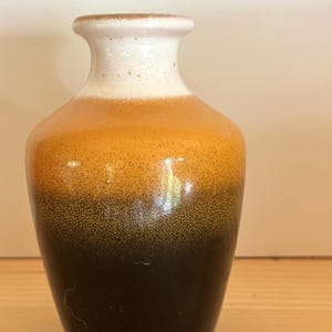 Könnte beinhalten: Eine kleine Keramikvase mit Farbverlauf. Die Oberseite ist weiß, geht in Orange über und dann in einen dunkelbraunen Sockel. Die Vase hat einen abgerundeten Körper und einen schmalen Hals, geeignet für kleine Blumen oder als dekoratives Stück.