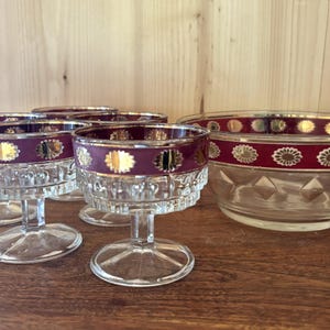Può includere: Un set di vetreria vintage, tra cui bicchieri da dessert con stelo e una ciotola da portata. I bordi sono decorati con una fascia bordeaux e motivi floreali e a pois dorati. I bicchieri e la ciotola sono trasparenti.