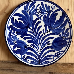 Op de afbeelding: Een ronde keramische bord met een witte ondergrond en handgeschilderde kobaltblauwe bloemmotieven. Het ontwerp bestaat uit grote, gestileerde blauwe bloemen en bladerrijke stengels in een symmetrisch patroon, afgewerkt met een blauwe rand.