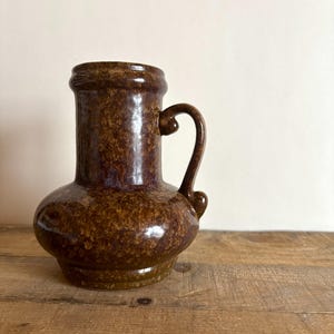 Può includere: Una brocca in ceramica marrone con una finitura smaltata maculata e screziata. Il recipiente presenta una base ampia e arrotondata che si restringe verso un collo cilindrico con un bordo svasato. Un manico curvo è fissato sul lato del corpo.