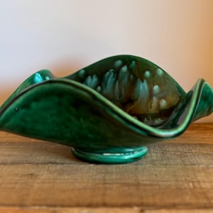 Könnte beinhalten: Eine Vintage-Keramikschale in Smaragdgrün mit einem einzigartigen, wellenförmigen Rand. Das Innere weist eine gesprenkelte, braune und cremefarbene Glasur auf, die dem Design Tiefe verleiht. Die Schale steht auf einem kleinen, kreisförmigen Sockel.