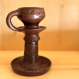 Könnte beinhalten: Ein handgefertigter, dunkelbrauner Kerzenhalter aus Keramik mit Griff. Der Kerzenhalter hat einen becherartigen Aufsatz mit kreisförmigen Designs, einen zentralen Stiel mit floralen und seilartigen Details und eine breite Basis. Er ist für eine Kerze konzipiert.