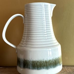 Könnte beinhalten: Eine weiße Keramikkrug mit einem gebogenen Griff und einem Ausguss. Der Krug hat horizontale graue Streifen in der Nähe der Oberseite und ein grünes und braunes Band am Boden. Der Krug ist etwa 25 cm hoch.