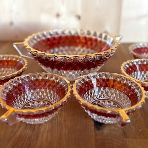 Könnte beinhalten: Ein Set aus sechs Vintage-Glasschüsseln. Die Schalen haben ein klares, rautenförmiges Muster mit einem roten Band und goldenen Verzierungen. Das Set enthält eine große Servierschüssel und fünf kleinere Schalen mit Griffen.