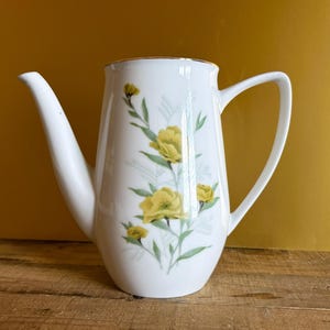Könnte beinhalten: Eine weiße Kaffeekanne aus Porzellan mit einer zarten botanischen Illustration gelber Blüten mit grünen Stielen und Blättern. Das Gefäß hat einen hohen, zylindrischen Körper, einen geschwungenen Ausguss und einen Henkel mit goldenem Rand.