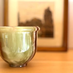 Può includere: Una tazza traslucida in onice verde con venature marroni. La tazza ha una forma arrotondata con un bordo svasato. Lo sfondo presenta un'immagine sfocata incorniciata. La tazza è appoggiata su una superficie di legno.