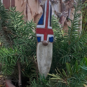 May include: A ceramic gnome figurine with a white beard and a hat painted with the colors of the Icelandic flag. The gnome is standing in a pot of green plants.