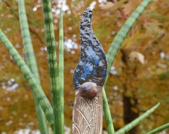 May include: A ceramic garden stake with a blue and brown glaze. The stake is shaped like a flame and has a textured surface. The stake is inserted into a pot of green plants.