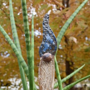 May include: A ceramic garden stake with a blue and brown glaze. The stake is shaped like a flame and has a textured surface. The stake is inserted into a pot of green plants.