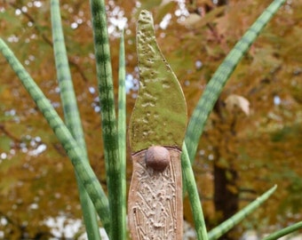 May include: A ceramic garden gnome stake with a green hat and a brown body. The gnome is standing in a pot of green plants.